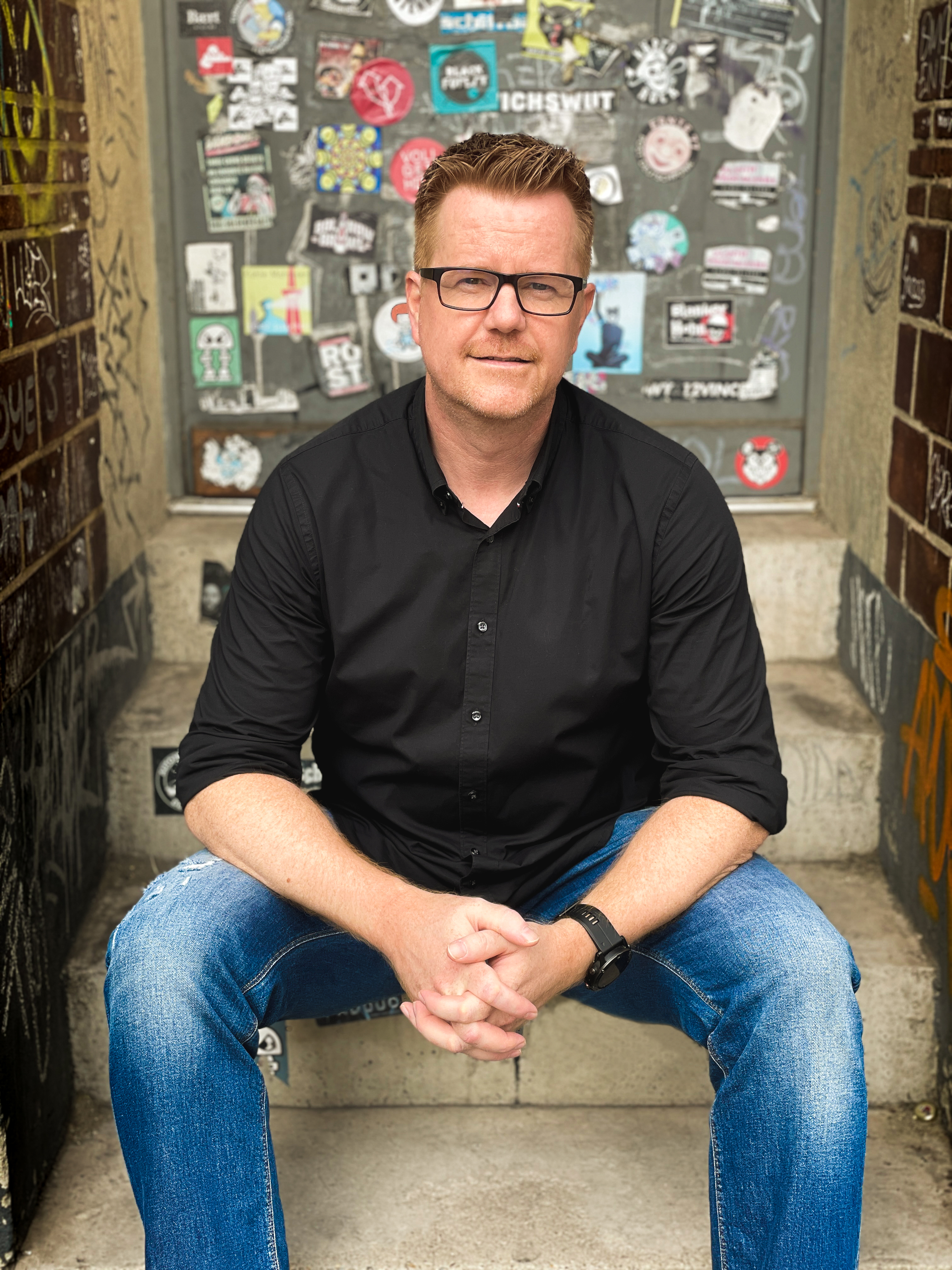 Zu sehen ist Friedrich Kalpenstein auf einer Treppe sitzend. Hinter ihm ist eine Tür mit vielen Aufklebern beklebt. Er trägt ein schwarzes Hemd, eine Jeans und eine Brille.